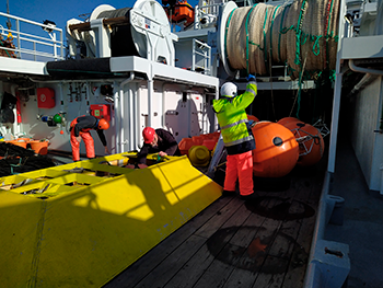 Fortøjningsarbejde på R/V Jákup Sverri i maj 2021. Alle strømmålere er hentet op og ligger på dækket. Der gøres klar til transport til Havstovan, hvor data læses og friske batterier sættes i til nye udlægninger. Forrest en strømmåler i trawlsikret ramme (gul), som har været udlagt på Island-Færø ryggen og bagved de andre strømmålere i orange flotation bøjer ( Karin M. H. Larsen).